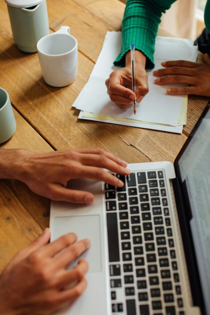 Two people working at a timber table, one typing on a laptop and the other writing notes
