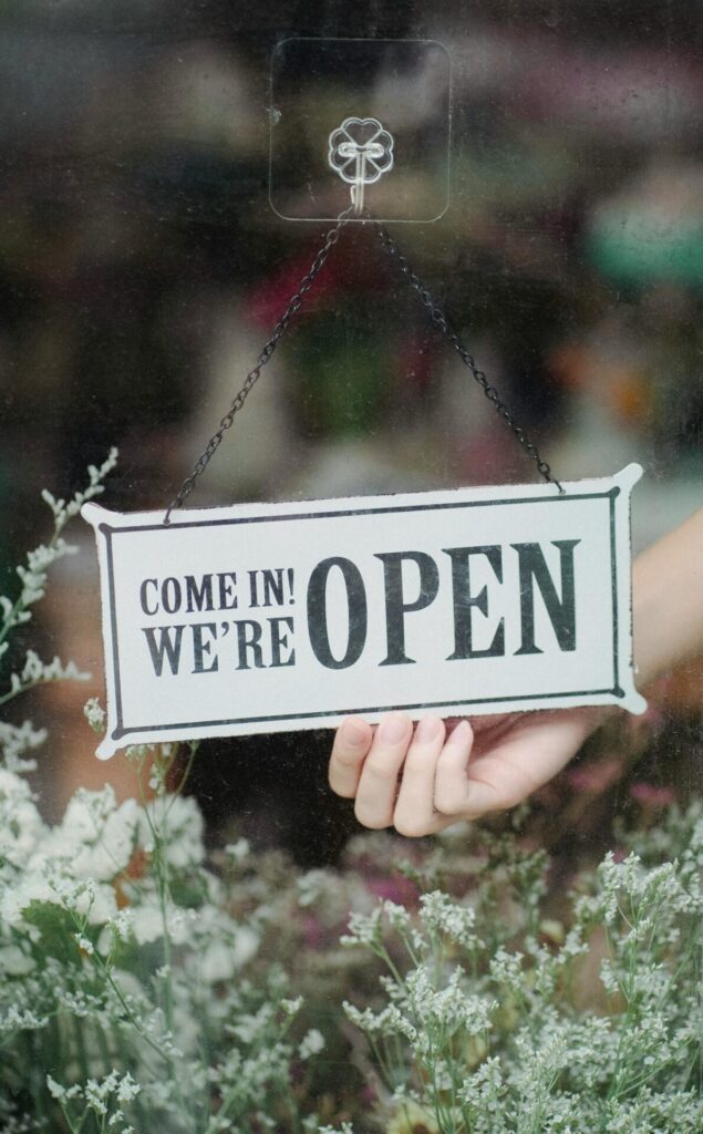 Hand turning a Come In We're Open sign in a shop window surrounded by flowers